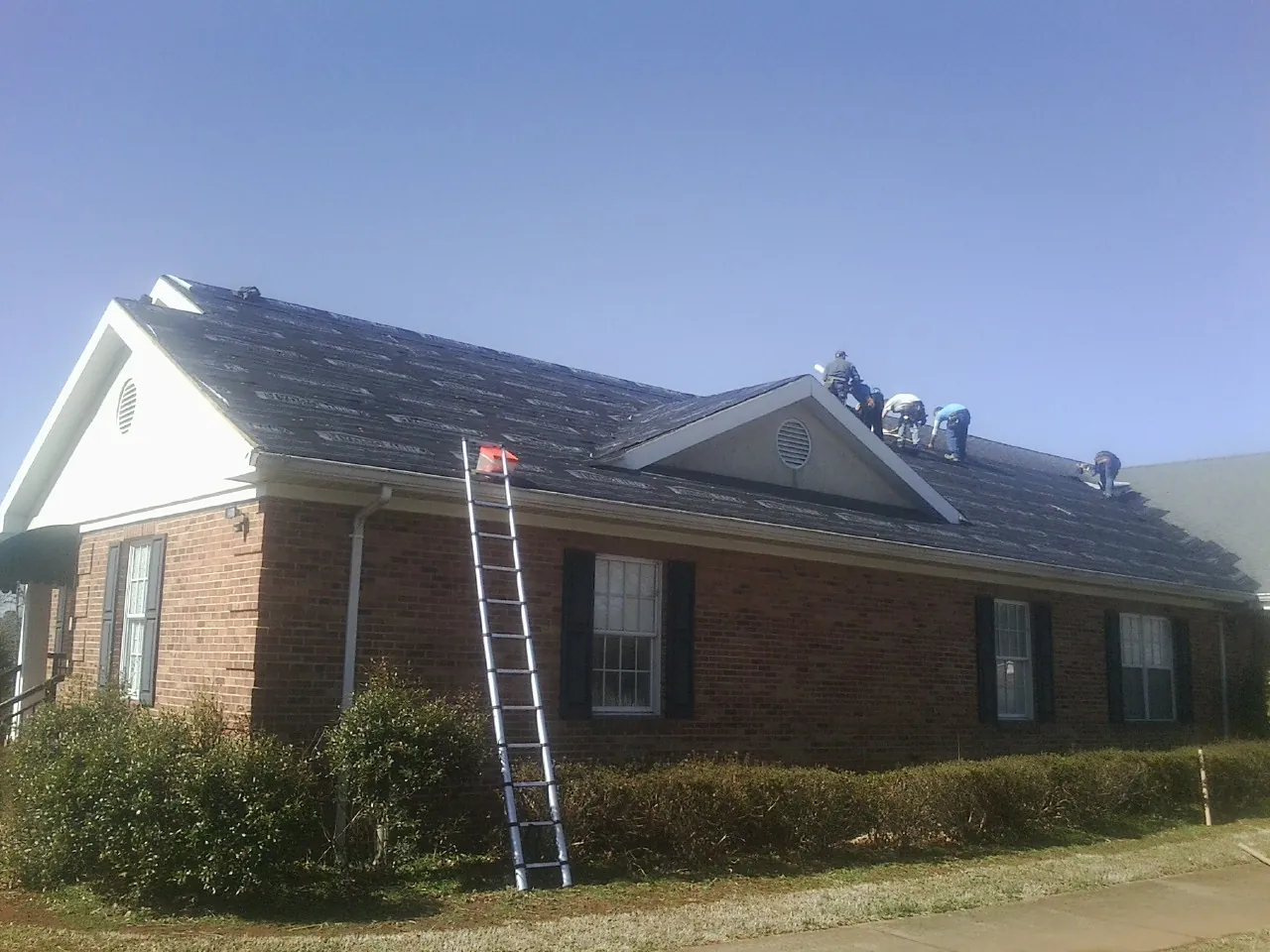 Crew tearing shingles off a brick ranch roof with a ladder set — Metal Roof Repair work by our crew in Meredith